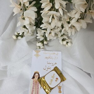 Puede incluir: Una tarjeta blanca con texto &aacute;rabe y una ilustraci&oacute;n de una mujer con vestimenta tradicional, acompa&ntilde;ada de un art&iacute;culo dorado con la inscripci&oacute;n "With Love". Flores blancas adornan la escena.