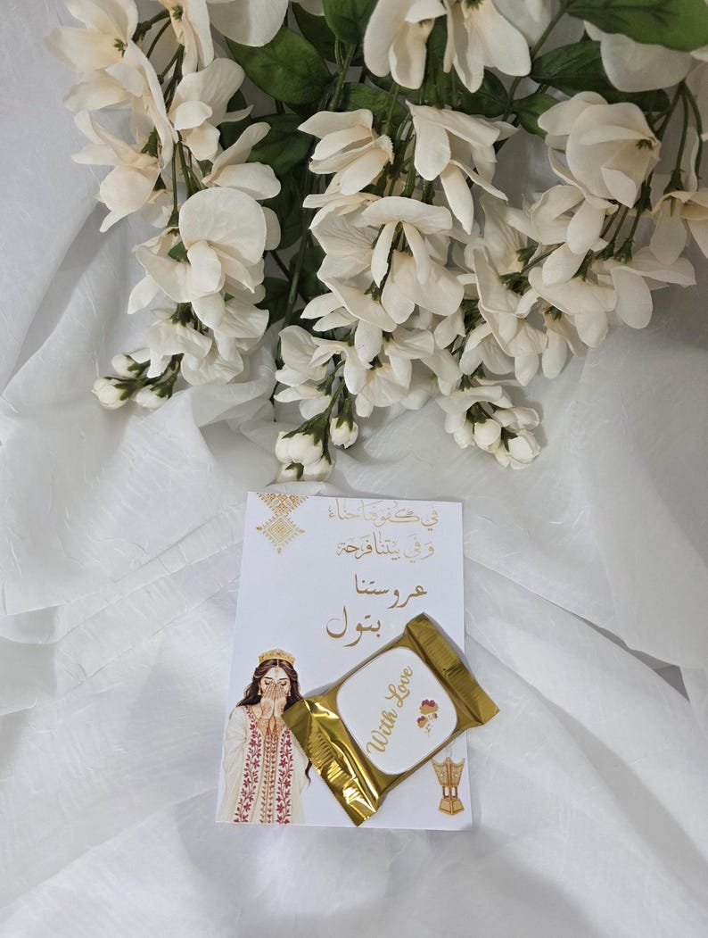Puede incluir: Una tarjeta blanca con texto &aacute;rabe y una ilustraci&oacute;n de una mujer con vestimenta tradicional, acompa&ntilde;ada de un art&iacute;culo dorado con la inscripci&oacute;n "With Love". Flores blancas adornan la escena.