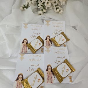 Puede incluir: Cuatro tarjetas blancas con texto e ilustraciones doradas, cada una combinada con un chocolate envuelto en oro. Las tarjetas presentan a una mujer con vestimenta tradicional y texto &aacute;rabe. Los chocolates tienen impreso "With Love". Flores blancas en el fondo.