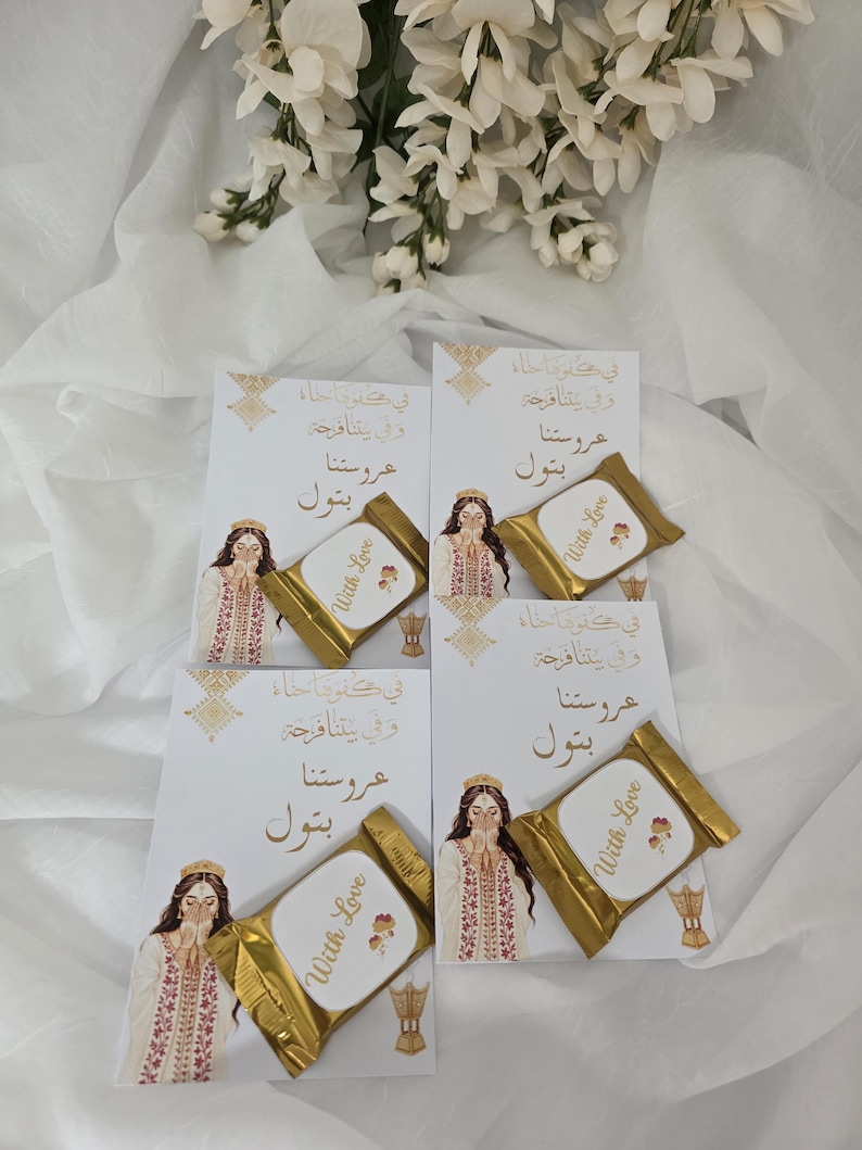 Puede incluir: Cuatro tarjetas blancas con texto e ilustraciones doradas, cada una combinada con un chocolate envuelto en oro. Las tarjetas presentan a una mujer con vestimenta tradicional y texto &aacute;rabe. Los chocolates tienen impreso "With Love". Flores blancas en el fondo.
