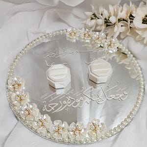 May include: Oval silver wedding ring tray with pearl and floral embellishments. Two white ring boxes sit on the tray. The tray has Arabic script and is surrounded by a pearl border and white flowers.