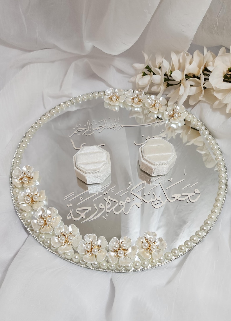 May include: Oval silver wedding ring tray with pearl and floral embellishments. Two white ring boxes sit on the tray. The tray has Arabic script and is surrounded by a pearl border and white flowers.