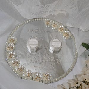 May include: A silver wedding ring tray with pearl and floral embellishments. The tray has two ring boxes and engraved Arabic script. The date 9.4.2025 is also engraved on the tray.