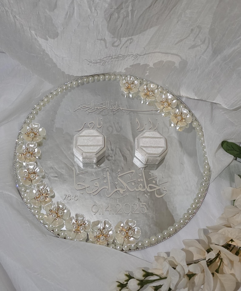 May include: A silver wedding ring tray with pearl and floral embellishments. The tray has two ring boxes and engraved Arabic script. The date 9.4.2025 is also engraved on the tray.