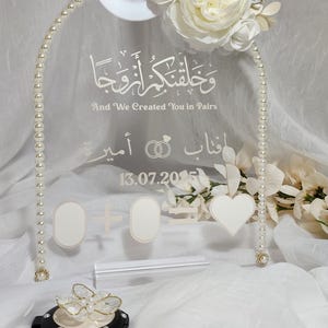 Op de afbeelding: Een heldere acryl bruiloftdecoratie met een parelboog, versierd met witte bloemen. De boog heeft Arabische tekst en Engelse tekst, samen met een datum en ringen. Inclusief een hart en twee ovale vormen.