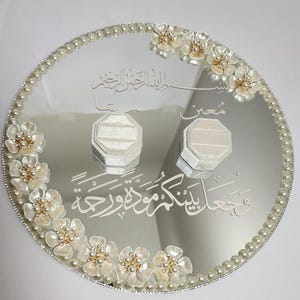 May include: A mirrored, round wedding ring tray with a pearl border and floral accents. Two ring boxes sit on the reflective surface, with Arabic script etched into the mirror. The flowers have pearl centers.