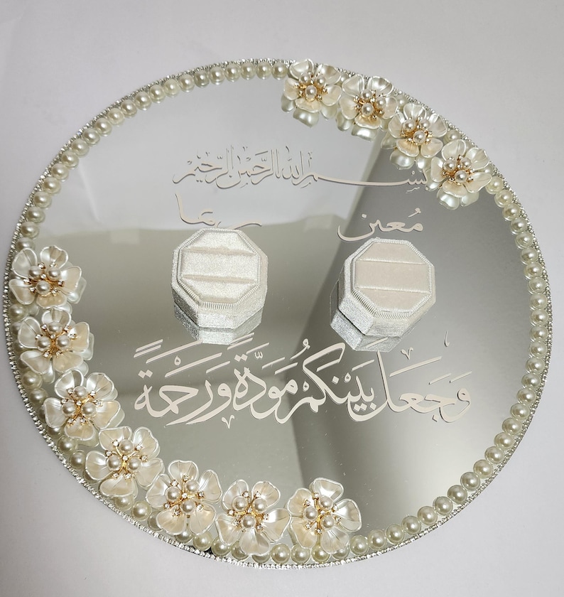 May include: A mirrored, round wedding ring tray with a pearl border and floral accents. Two ring boxes sit on the reflective surface, with Arabic script etched into the mirror. The flowers have pearl centers.