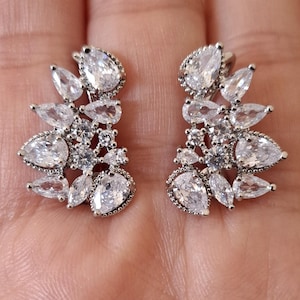 Puede incluir: Un par de pendientes de plata con un racimo de piedras de circonita cúbica transparentes en forma de lágrima. Los pendientes están diseñados para abrazar el lóbulo de la oreja y tienen un aspecto delicado y femenino.