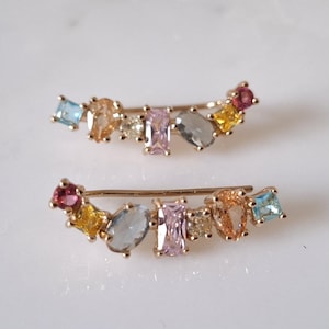 May include: A pair of gold ear cuffs with a curved design. Each cuff is adorned with a row of colourful gemstones, including pink, blue, yellow, and orange.