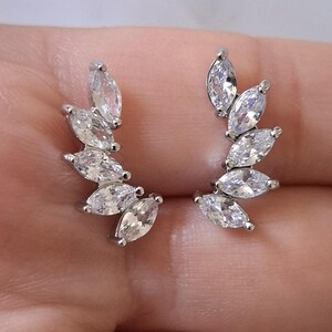 May include: A pair of silver ear climbers. Each climber has a series of marquise-cut, clear cubic zirconia stones, arranged in a curved, leaf-like design. The earrings are set against a neutral background.