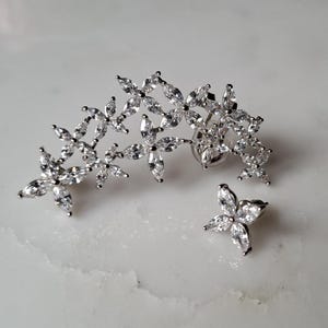 May include: A pair of silver earrings with a floral design. The larger earring is a long, curved piece with multiple flower shapes set with clear cubic zirconia stones. The smaller earring is a single flower shape with clear cubic zirconia stones.