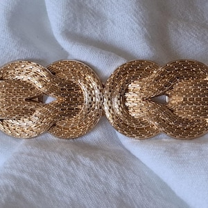 May include: A gold-coloured belt buckle with an intricate woven design. The buckle features two connected circular shapes, each with a textured surface, and a rectangular clasp. The buckle is set against a white fabric.