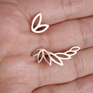 Puede incluir: Dos pendientes dorados. Uno es un pequeño pendiente de botón con un diseño de tres pétalos. El otro es un trepador de oreja más largo con una serie de formas de hojas caladas conectadas. Los pendientes se muestran sobre un fondo color piel.