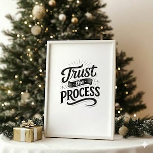 Peut inclure: Une affiche encadrée blanche avec l'inscription noire "Trust the Process". L'affiche est placée devant un sapin de Noël orné de boules et de guirlandes lumineuses dorées. Un coffret cadeau emballé est au premier plan.