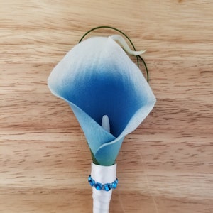 May include: A blue and white artificial calla lily boutonniere with a white satin ribbon and blue rhinestones.