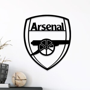 May include: Black metal Arsenal logo wall art. The design features the word "Arsenal" above a cannon emblem within a shield shape. The artwork is mounted on a white wall.
