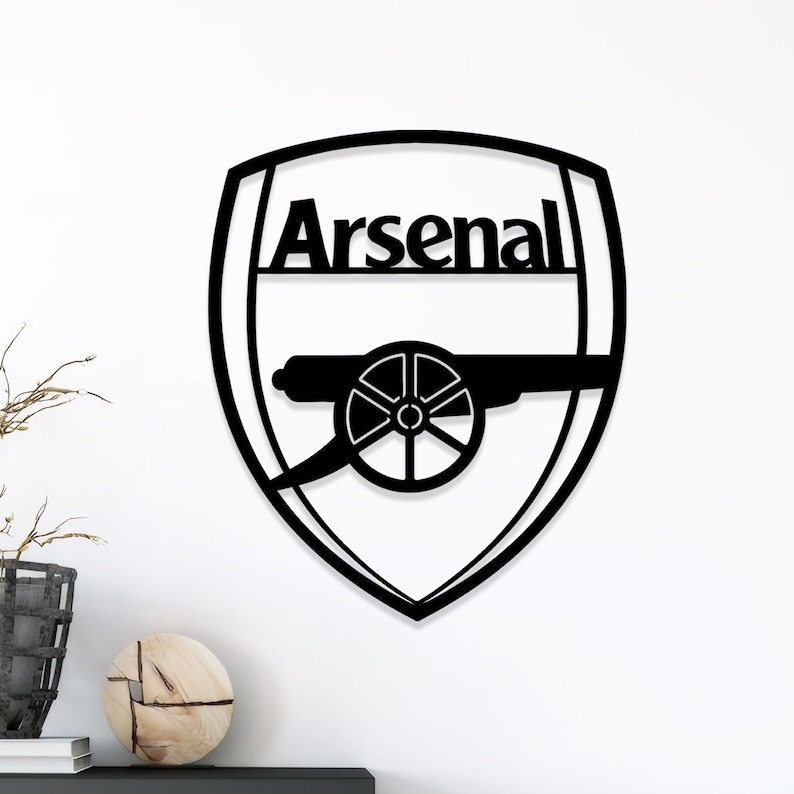 May include: Black metal Arsenal logo wall art. The design features the word "Arsenal" above a cannon emblem within a shield shape. The artwork is mounted on a white wall.
