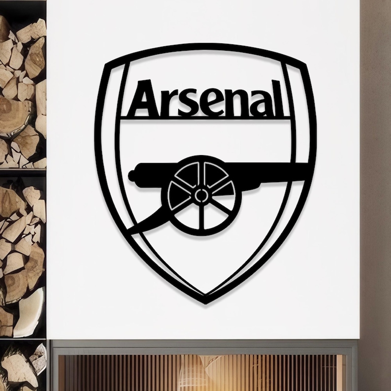 May include: Black metal Arsenal logo in a shield shape. The logo features the word "Arsenal" at the top, with a cannon in the center. The design is a bold, minimalist style, suitable for wall decor or fan merchandise.