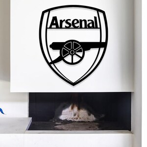 May include: Black metal wall art featuring the Arsenal football club logo. The logo is a shield shape with the word "Arsenal" at the top and a cannon in the center. The art is mounted on a white wall above a fireplace.
