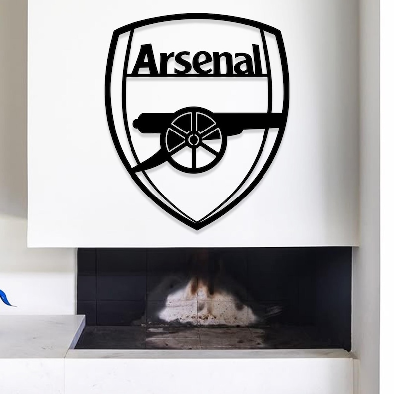 May include: Black metal wall art featuring the Arsenal football club logo. The logo is a shield shape with the word "Arsenal" at the top and a cannon in the center. The art is mounted on a white wall above a fireplace.