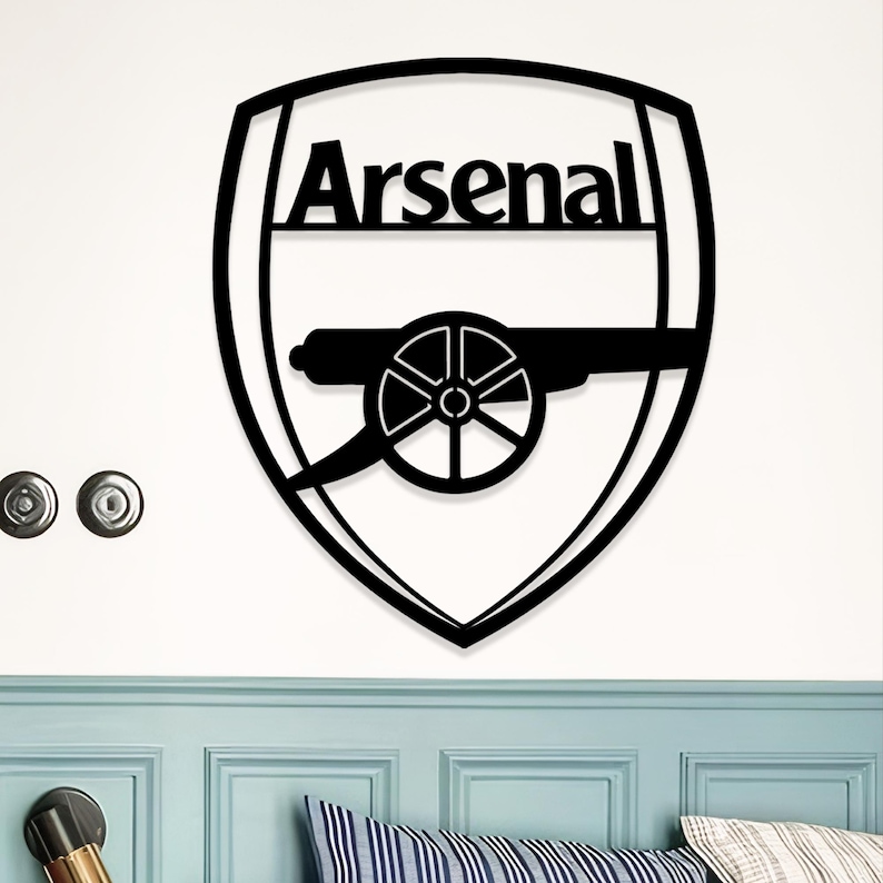May include: Black metal Arsenal logo wall art. The design features the word "Arsenal" above a cannon emblem, all within a shield shape. The artwork is mounted on a white wall.