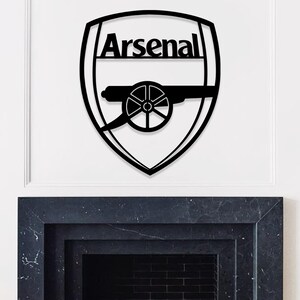 May include: Black metal wall art featuring the Arsenal logo. The logo is a shield shape with the word "Arsenal" at the top. Below the word is a cannon design. The art is mounted on a white wall.