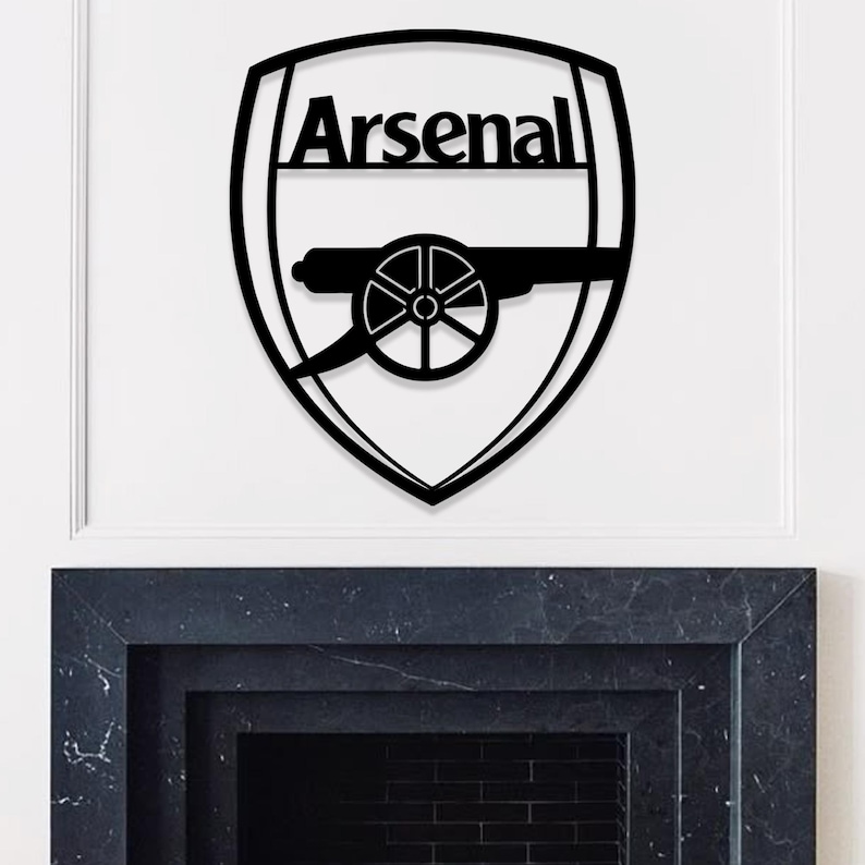 May include: Black metal wall art featuring the Arsenal logo. The logo is a shield shape with the word "Arsenal" at the top. Below the word is a cannon design. The art is mounted on a white wall.