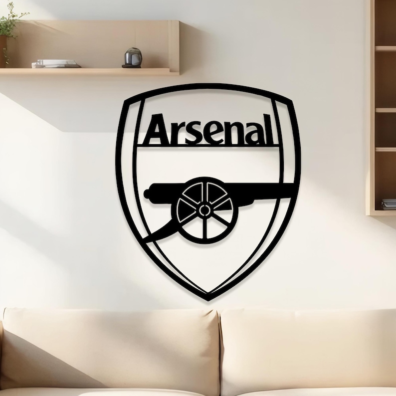 May include: Black metal wall art featuring the Arsenal logo. The logo includes the word "Arsenal" above a cannon emblem, all within a shield shape. The artwork is mounted on a white wall above a beige sofa.