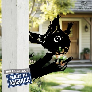 May include: Black metal squirrel yard art. The squirrel is peeking around a white post and giving the middle finger. The squirrel has a detailed design and is suitable for outdoor use.
