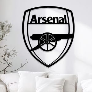May include: Black metal wall art featuring the Arsenal logo. The logo is a shield shape with the word "Arsenal" at the top and a cannon in the center. The art is mounted on a white wall above a white couch.