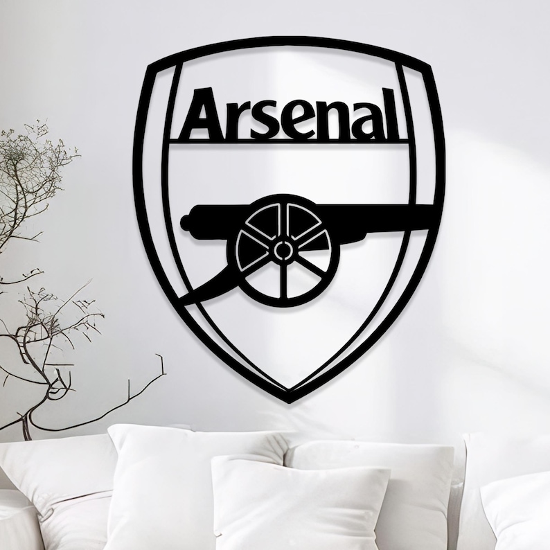 May include: Black metal wall art featuring the Arsenal logo. The logo is a shield shape with the word "Arsenal" at the top and a cannon in the center. The art is mounted on a white wall above a white couch.