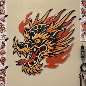May include: A wall-mounted wooden cutout of a dragon head featuring a vibrant color palette of yellow, orange, and black. The dragon is depicted with an open mouth, sharp teeth, and flowing flame-like details extending from the back of its head.