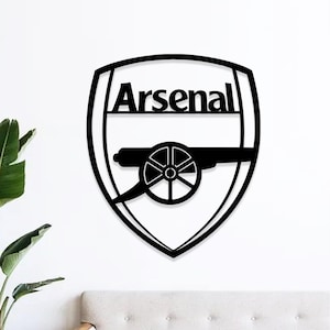 May include: Black metal wall art featuring the Arsenal logo. The design includes the word "Arsenal" above a cannon, all within a shield shape. The artwork is mounted on a white wall.