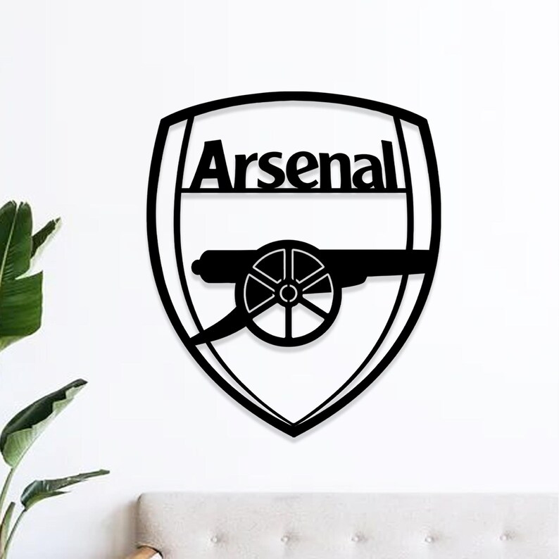 May include: Black metal wall art featuring the Arsenal logo. The design includes the word "Arsenal" above a cannon, all within a shield shape. The artwork is mounted on a white wall.