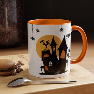 Pode incluir: Caneca de cerâmica branca com interior e alça laranja. A caneca apresenta uma silhueta preta de uma casa assombrada, uma lua cheia, aranhas e uma bruxa. Design temático de Halloween.