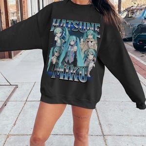 May include: Black sweatshirt featuring the text "Hatsune Miku" in a blue and white design. The sweatshirt has a graphic of the anime character Hatsune Miku in various poses. The sweatshirt is a crew neck style.