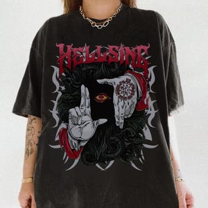 May include: Black t-shirt featuring the word "HELLSING" in red gothic lettering. The graphic includes a stylized illustration of hands framing an eye, with intricate tattoo details and a dark, textured background. The shirt has a vintage wash.