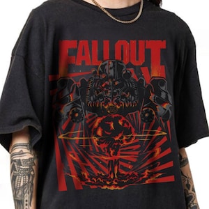 May include: A black short-sleeve t-shirt featuring a graphic print of a power-armored figure above a mushroom cloud. The text "FALLOUT" is printed in large red letters at the top. The design includes red sunburst lines radiating from the center.