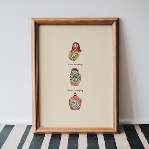 Puede incluir: Impresión artística enmarcada con tres muñecas rusas Matryoshka en acuarela. Las muñecas son rojas, verdes y amarillas, con los textos "Ever Growing" y "Ever Changing" debajo. El marco es de color marrón claro.