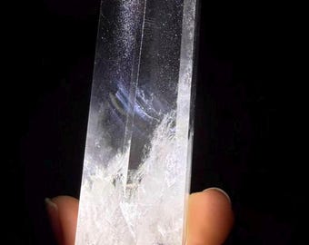 May include: A clear, faceted quartz crystal point with a hexagonal shape. The stone features a transparent body with internal white inclusions and natural fractures, catching the light to create subtle reflections against a dark background.