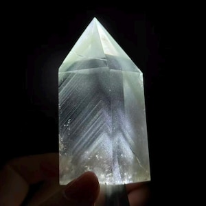 May include: A clear, faceted crystal point with a pointed top and a hexagonal base. The interior of the stone displays distinct, angular chevron-like patterns and internal fractures that catch the light, creating a bright, glowing effect.