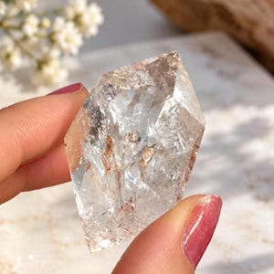 May include: A clear, faceted quartz crystal with internal fractures and subtle reddish-brown mineral inclusions. The stone has a geometric, multi-sided shape with sharp edges and a polished, reflective surface that catches the light.