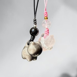 Puede incluir: Dos colgantes de piedras preciosas con forma de gato. Uno es de color gris metálico, suspendido de un cordón negro. El otro es de color rosa claro, colgado de un cordón rosa. Ambos tienen una cuenta encima del colgante de gato.