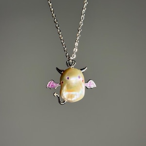 May include: A silver necklace featuring a unique pendant. The pendant is a stylized devil figure crafted from a lustrous, cream-colored pearl. It has small horns, purple eyes, and pink wings. The necklace chain is delicate and silver.