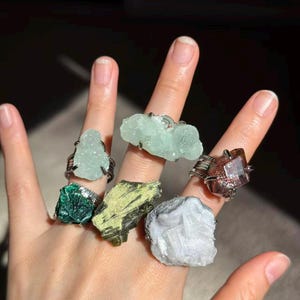 May include: A hand displays six rings, each featuring a different raw gemstone. The gemstones are various colors, including green, blue, and brown, set in silver-toned wire. The rings showcase the natural beauty of the crystals.