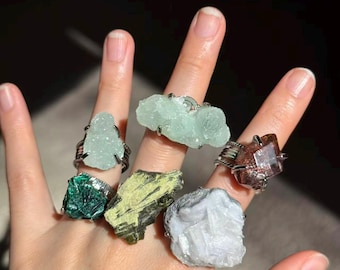 Könnte beinhalten: Eine Hand zeigt sechs Ringe, jeder mit einem anderen rohen Edelstein. Die Edelsteine sind in verschiedenen Farben, darunter Grün, Blau und Braun, in silberfarbenem Draht gefasst. Die Ringe zeigen die natürliche Schönheit der Kristalle.