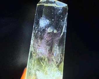 May include: A clear, faceted, rectangular crystal-like object illuminated from above by a bright light source. The interior contains suspended, wispy, purple and white inclusions that resemble delicate floral or botanical structures.