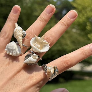 Könnte beinhalten: Eine Sammlung von Silberringen, jeder mit einem einzigartigen Muscheldesign. Die Ringe zeigen verschiedene Muscheln, darunter einen Seestern, eine Muschel und Jakobsmuscheln, einige mit Perlenakzenten. Die Ringe werden auf einer Hand präsentiert.