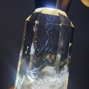Può includere: Un cristallo trasparente e sfaccettato con fratture e inclusioni interne. Il cristallo è illuminato dall'alto, evidenziando la sua forma geometrica e i dettagli interni. Il colore generale è giallo pallido con aree bianche e iridescenti.