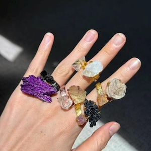 May include: A hand displaying a collection of gemstone rings. The rings feature various raw, uncut stones in colors like purple, black, clear, and gold, set on gold or silver bands. The rings are of different sizes and shapes.